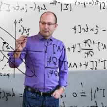 Christian Hesse steht vor einer Tafel aus Glas und schreibt mathematische Formeln darauf.